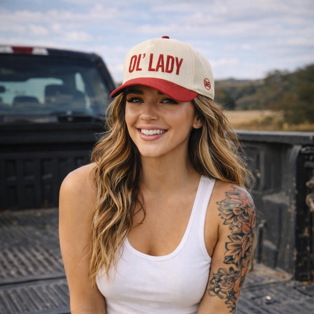 Woman wearing a 'OL' LADY' cap sitting on a truck bed with a scenic background